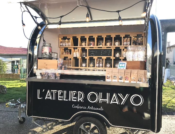 Remorque noire à l'enseigne L'Atelier Ohayo, auvent ouvert et guirlande allumée, présentoir de confiseries en bocaux
