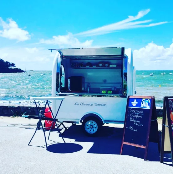 Remorque blanche ouverte face à la mer, ardoise annonçant boissons, gaufres et glaces